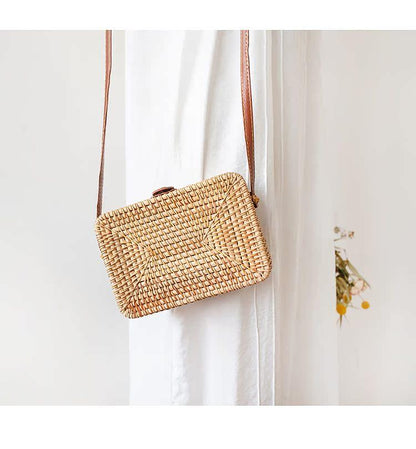 Stylish rattan box bag with natural woven design hanging on a white curtain, featuring a brown strap for casual outings.