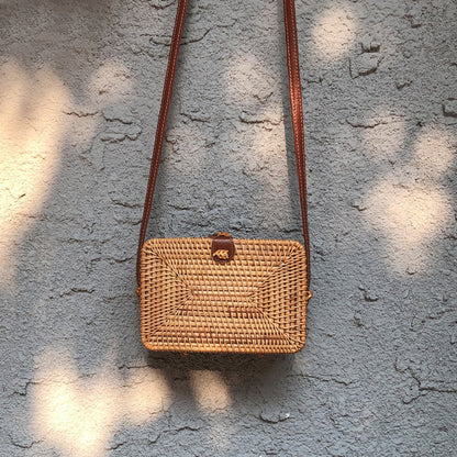 Rattan Box Bag with brown leather strap hanging against a textured wall